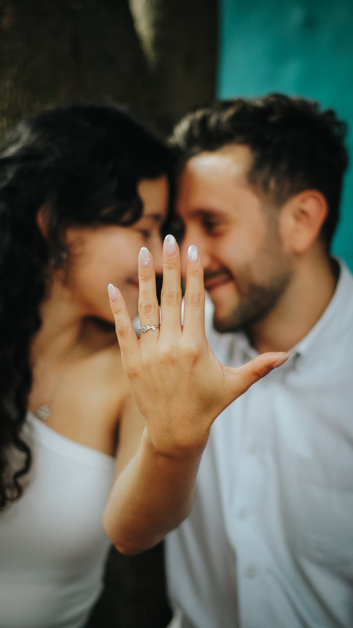 The engagement ring held up to the camera, the couple soft behind it