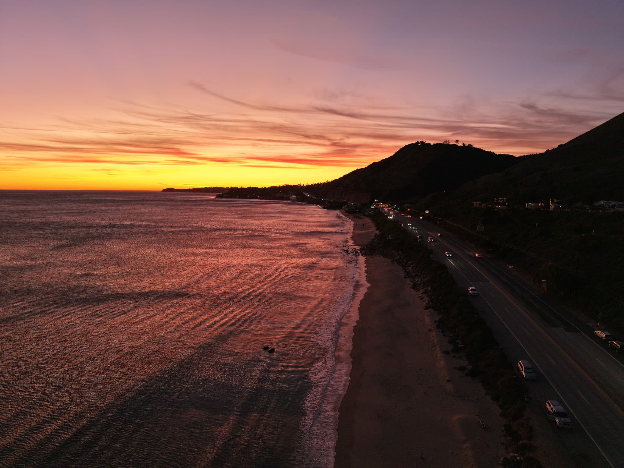 The coast road at dusk from above, surf on one side and headlights on the other