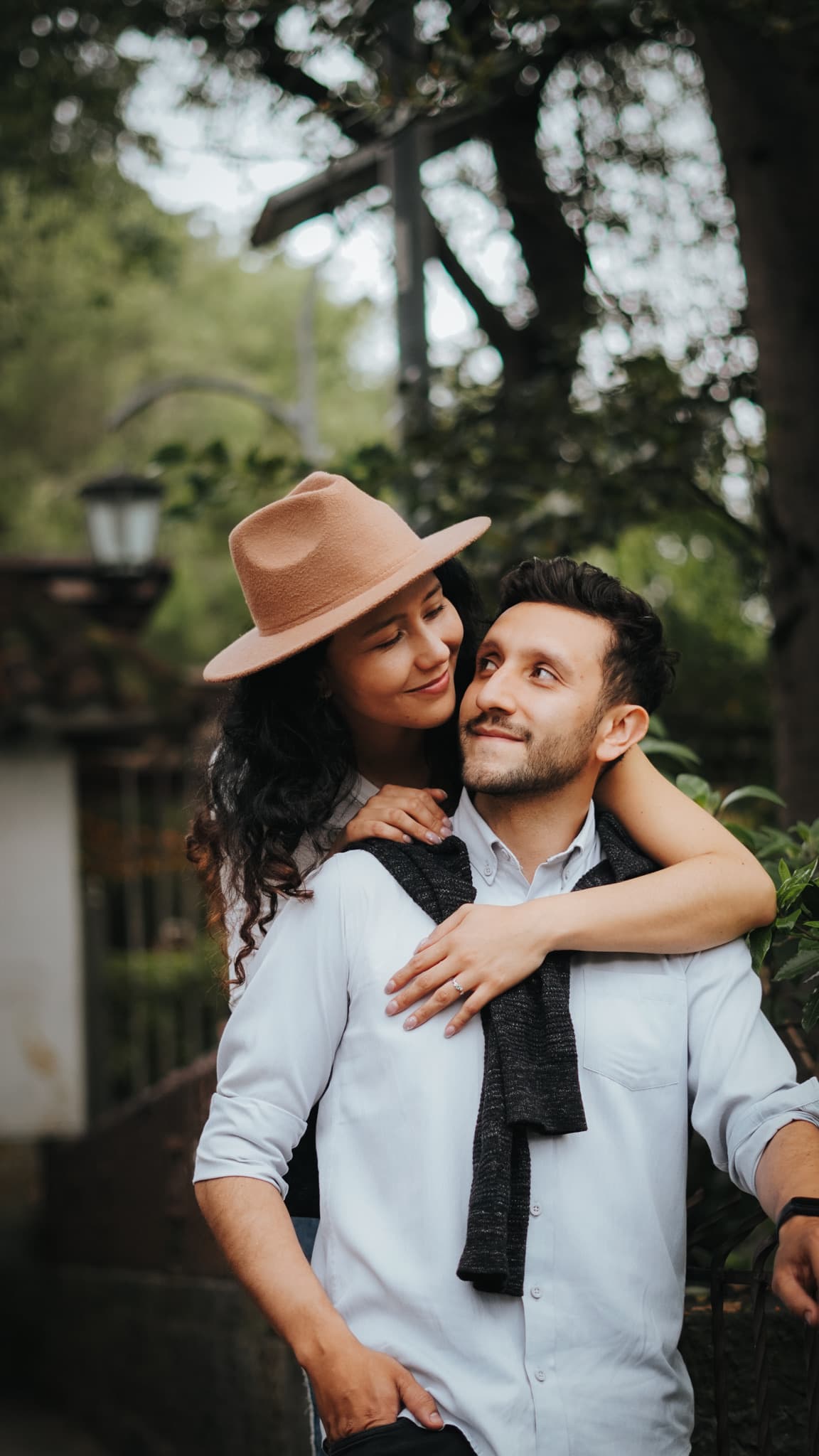 She leans over his shoulder at a garden gate, her ring hand on his chest