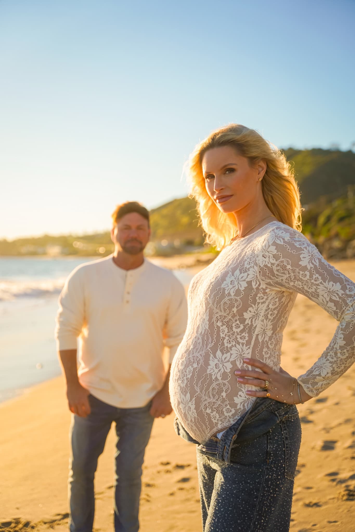 An expectant mother on the shoreline at golden hour, her partner behind her
